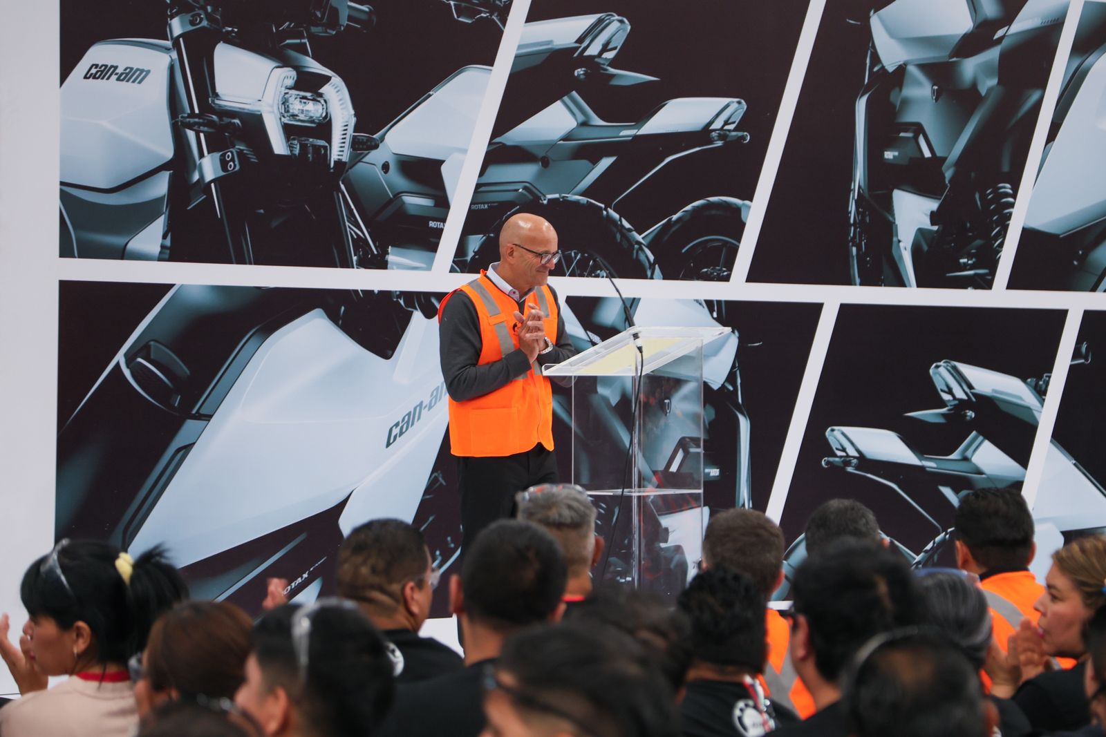 Executive in a safety vest speaking at a lectern in front of a wall of Can-Am electric motorcycle images, before plant employees